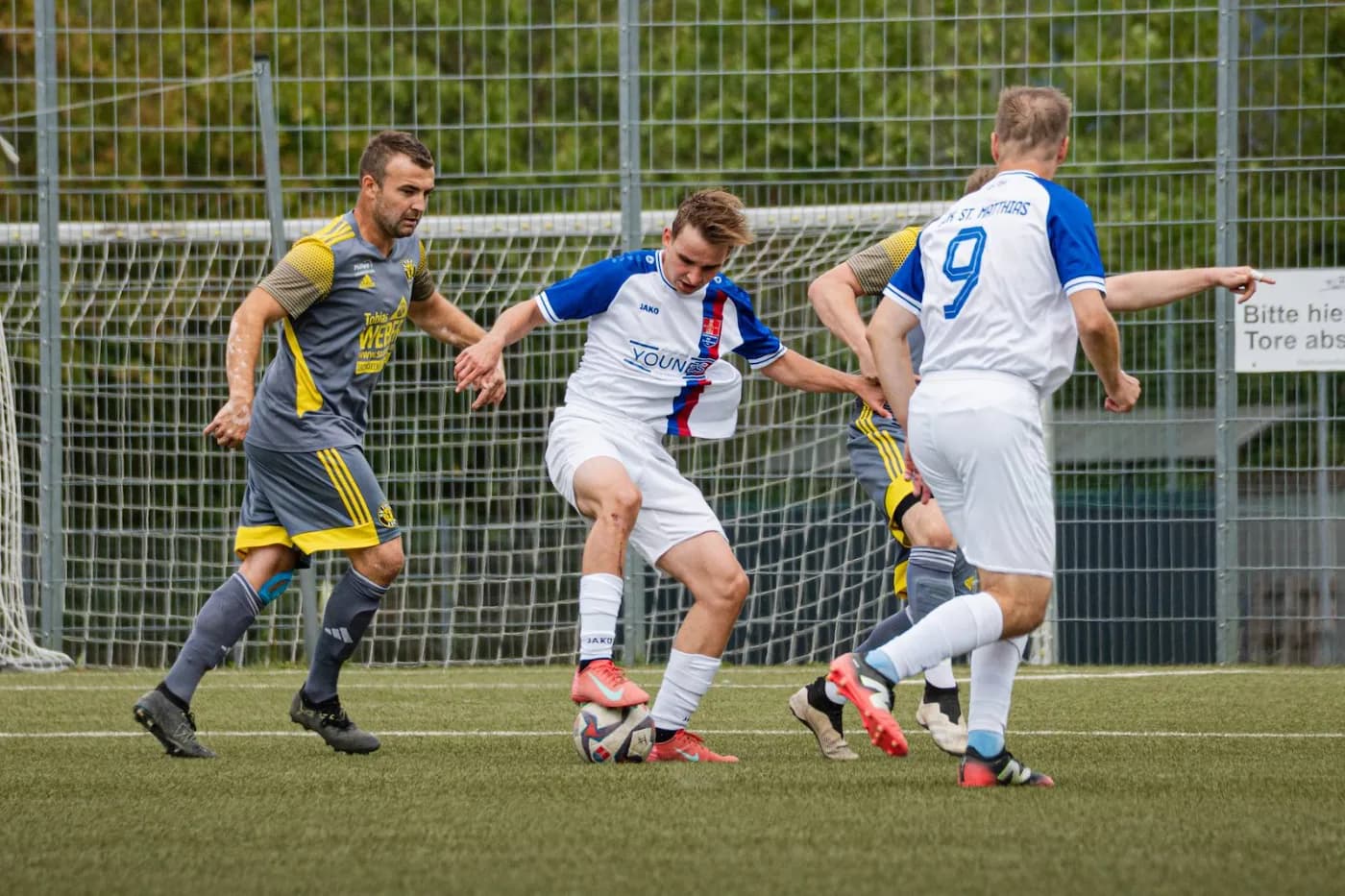 Drei Spieler kämpfen um den Ball, zwei im weißen Trikot des Gegners, einer in Grau-Gelb