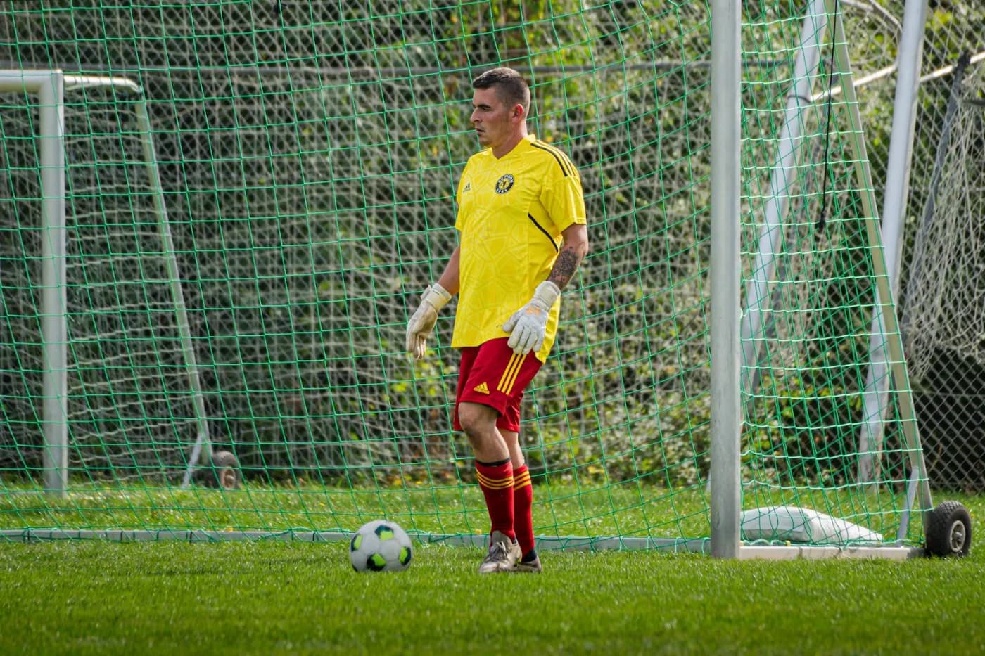 Ein Torwart im roten Trikot steht vor dem Tor, der Ball liegt im Gras