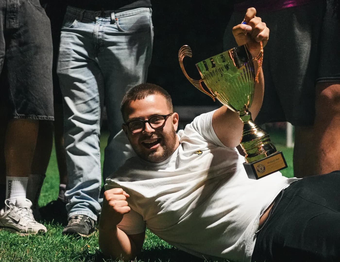 Ein Spieler des SV Fisch liegt lachend im Gras und streckt den Siegerpokal des Elferturniers in die Höhe